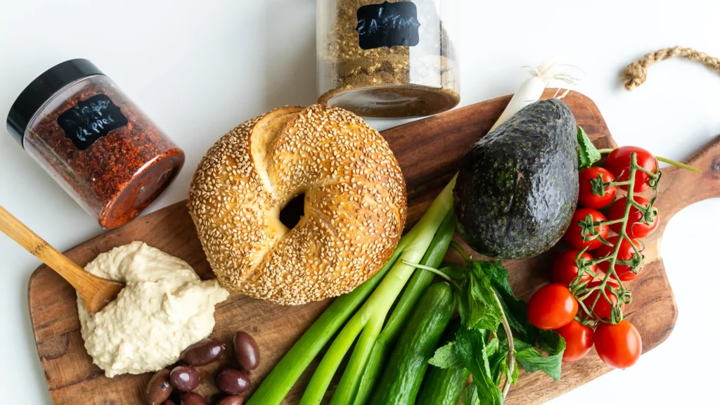 A bagel, avocado and other vegetables on a cutting board.