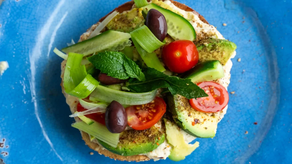 A bagel, avocado and other vegetables on a cutting board.