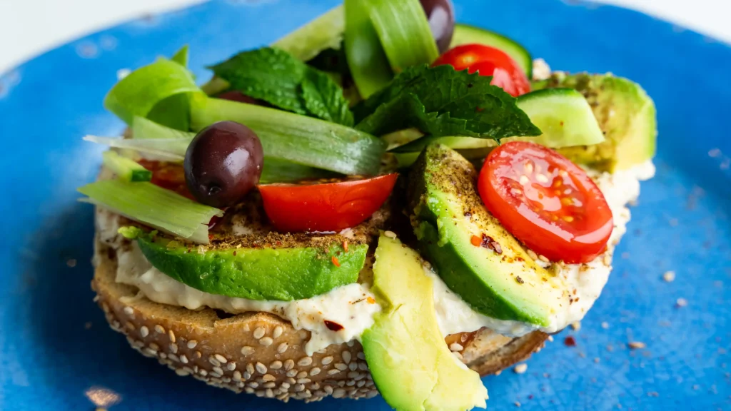 A bagel, avocado and other vegetables on a cutting board.